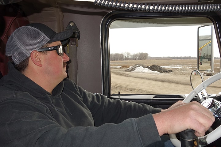 A truck driver wearing a hat gazes out of the truck's window, scanning the surroundings while on the road.